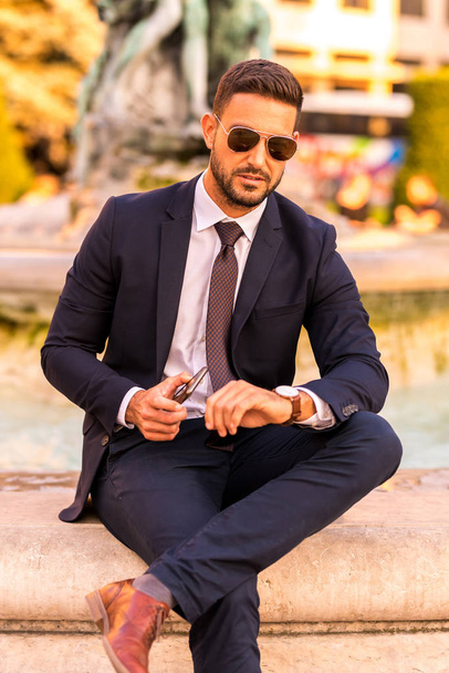 A businessman sitting next to a fountain and checking the time - 写真・画像