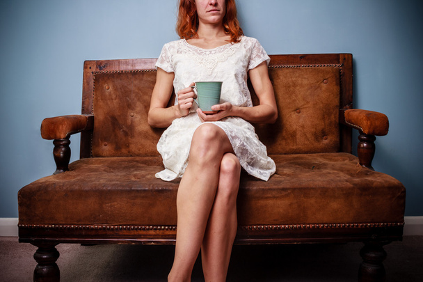 Young woman in summer dress sitting on old sofa drinking tea - Фото, изображение