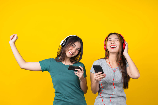 Две счастливые азиатские улыбающаяся молодая женщина в беспроводных наушниках для прослушивания музыки через смартфон и танцы на изолированном желтом фоне, стиль жизни и отдыха с хобби концепции
 - Фото, изображение