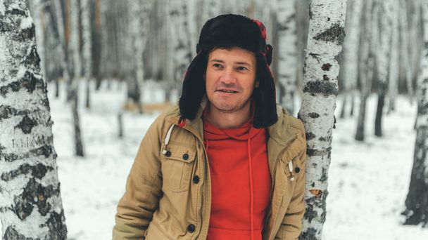Man standing in forest in winter day. Serious young male wearing beige jacket over red hoodie and earflap hat standing on snowy forest and looking away - Φωτογραφία, εικόνα