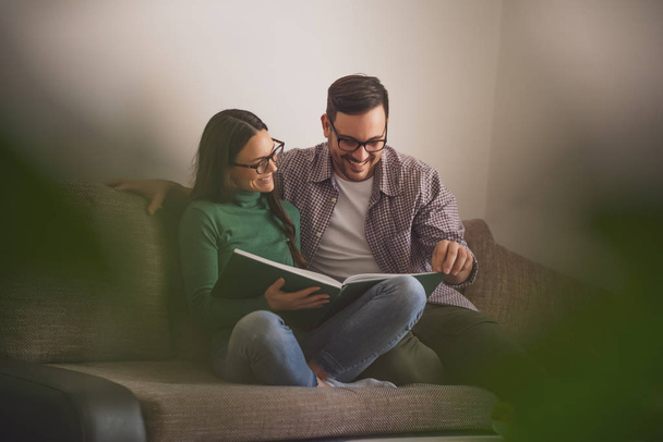 Happy couple is sitting at sofa in their home and looking at book. - Fotoğraf, Görsel