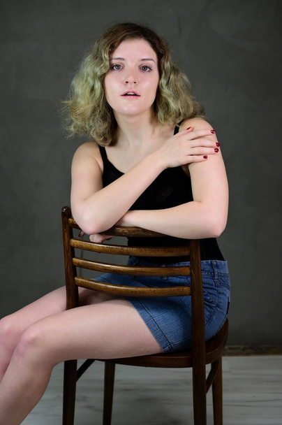 Portrait of a pretty student girl with beautiful curly hair in front of the camera on a gray background. Concept vertical photo of a young woman in a black T-shirt and blue skirt sitting on a chair. - Photo, image