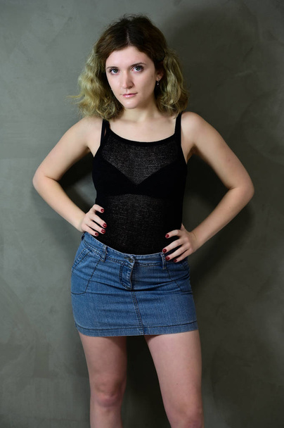 Concept vertical photo of a young woman in a black T-shirt and blue skirt standing straight. Portrait of a pretty student girl with beautiful curly hair in front of the camera on a gray background. - Zdjęcie, obraz