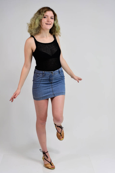 A horizontal photo concept of a young woman in a black t-shirt and blue skirt is standing in front of the camera on a white background. Full-length portrait of a pretty girl with beautiful curly hair. - Foto, imagen