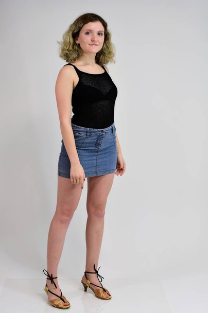A horizontal photo concept of a young woman in a black t-shirt and blue skirt is standing in front of the camera on a white background. Full-length portrait of a pretty girl with beautiful curly hair. - Foto, imagen