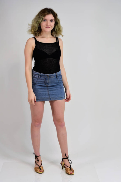 Full-length portrait of a pretty girl with beautiful curly hair. A horizontal photo concept of a young woman in a black t-shirt and blue skirt is standing in front of the camera on a white background. - Fotografie, Obrázek
