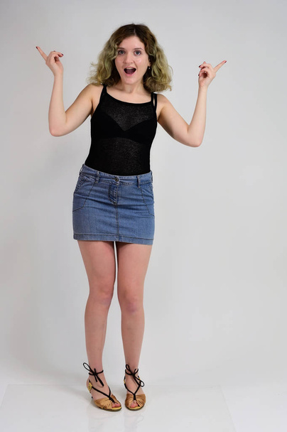 Full-length portrait of a pretty girl with beautiful curly hair. A horizontal photo concept of a young woman in a black t-shirt and blue skirt is standing in front of the camera on a white background. - Фото, зображення