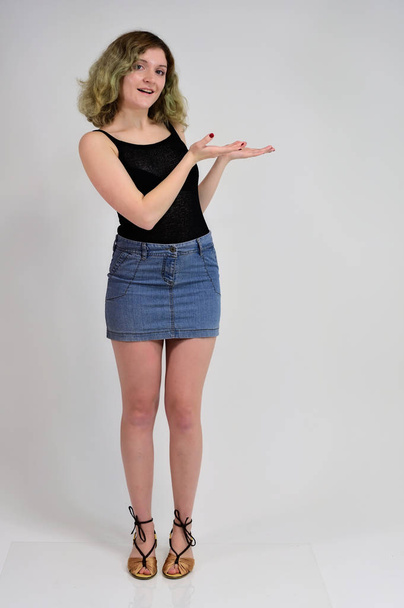 Full-length portrait of a pretty girl with beautiful curly hair. A horizontal photo concept of a young woman in a black t-shirt and blue skirt is standing in front of the camera on a white background. - 写真・画像