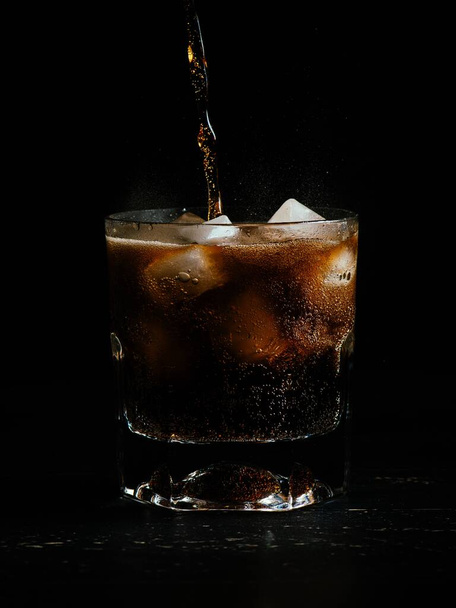 Vertical shot of coke being poured in a glass full of ice on a black background - Фото, изображение