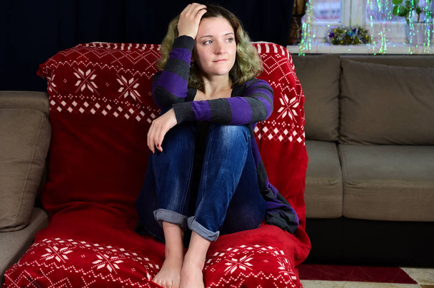 Portrait of a girl with beautiful magnificent hair rejoices on a sofa. Horizontal concept photo of a young woman in a sweater and jeans sits on a chair in front of the camera in the home interior. - Foto, Imagem