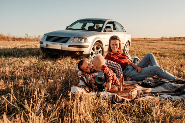 Happy Young Family Mom and Dad with Their Little Son Enjoying Summer Weekend Picnic Sitting on the Plaid Near the Car Outside the City in the Field at Sunny Day Sunset, Vacation and Road Trip Concept - Valokuva, kuva