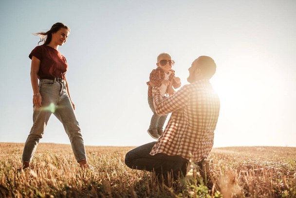 Gelukkig jong gezin mam en pap met hun kleine zoon genieten van de zomer weekend picknick buiten de stad in het veld op zonnige dag zonsondergang, vakantie tijd concept - Foto, afbeelding