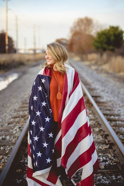Vertical shot of a female with the American flag on her shoulders standing on the railway - Photo, Image