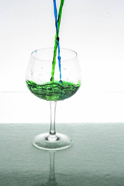 A vertical shot of green and blue liquid pouring in a glass cup with a white background - Фото, изображение