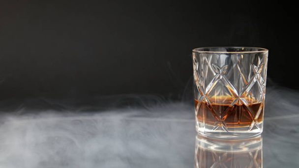 Glass of whiskey on a table surrounded by smoke against a black background - Foto, afbeelding