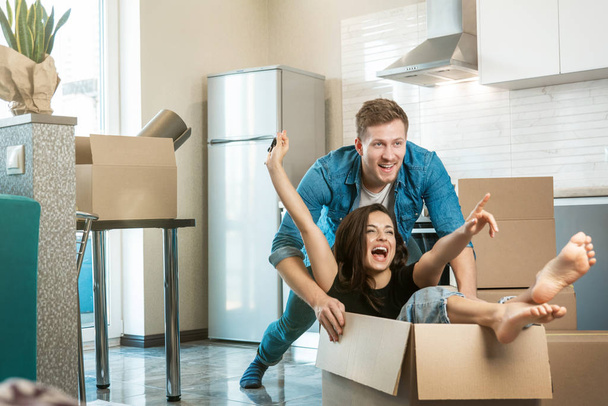 Genç çift kutuları açarken ve yeni bir daireye taşınırken eğleniyor. Koca, karısını kutuda yerde taşıyor. - Fotoğraf, Görsel