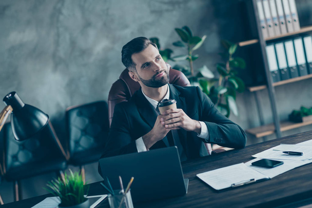 Photo of successful business guy hold takeout hot coffee beverage mug thoughtfully look up enjoy pleasant drink wear black blazer shirt tie suit costume sit chair office indoors - Photo, Image