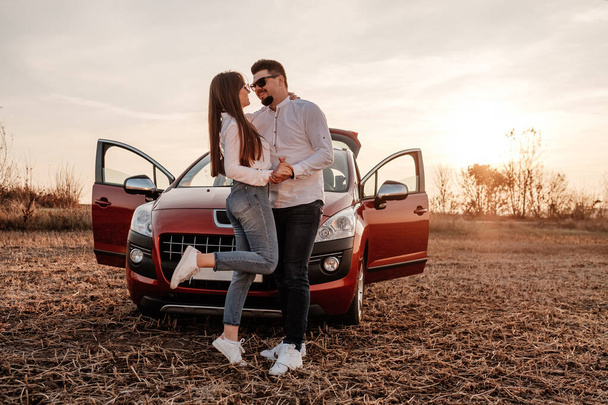 Young Happy Couple Dressed Alike in White Shirt and Jeans Enjoying Road Trip at Their New Car, Beautiful Sunset on the Field, Vacation and Travel Concept - Foto, imagen