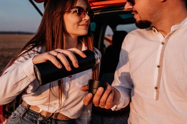 Giovane coppia felice vestita allo stesso modo in camicia bianca e jeans godendo di viaggio su strada alla loro nuova auto, bellissimo tramonto sul campo, vacanza e concetto di viaggio
 - Foto, immagini