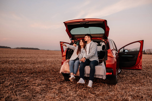 白いシャツを着た若い幸せなカップルと新しい車のトランクに座っているジーンズ、フィールドでの美しい夕日、休暇と旅行のコンセプト - 写真・画像