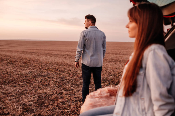 Giovane coppia felice vestita allo stesso modo in camicia bianca e jeans seduti al loro nuovo bagagliaio auto, bellissimo tramonto sul campo, vacanza e concetto di viaggio
 - Foto, immagini
