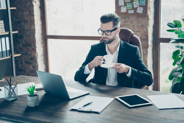 Photo of handsome business chief guy holding hot coffee beverage mug looking notebook screen talking skype relatives wear specs black blazer shirt suit sit chair office indoors - Fotoğraf, Görsel