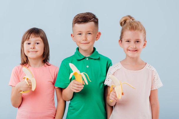 caucasian three kids with bananas in hand. isolated on blue background, copy space, in studio - Foto, Bild