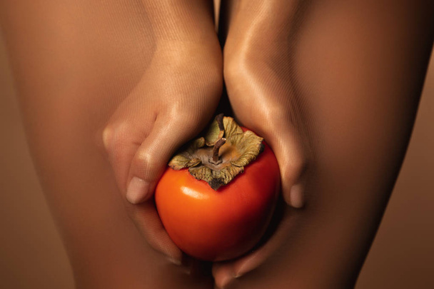 cropped view of woman in nylon tights holding ripe persimmon isolated on brown - Foto, Imagem