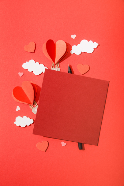 top view of paper heart shaped air balloons in clouds near blank card and pencil on red background - Foto, Imagen