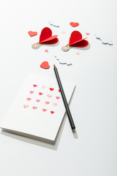 selective focus of greeting card with hearts and pencil near paper heart shaped air balloons in clouds on white background - Fotó, kép