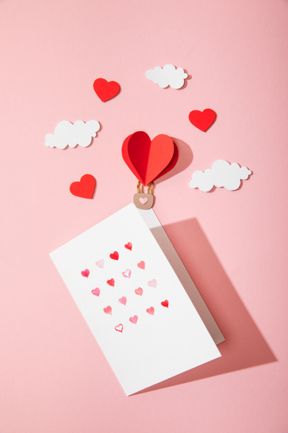 top view of greeting card with hearts in white envelope near paper heart shaped air balloon in clouds on pink - Photo, Image