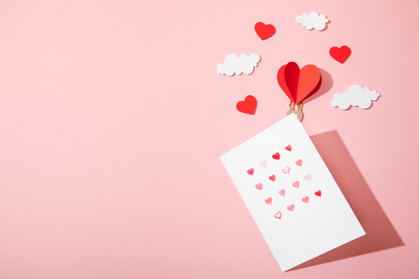 top view of greeting card with hearts in white envelope near paper heart shaped air balloon in clouds on pink - Zdjęcie, obraz