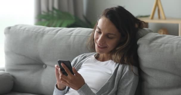 Happy young woman relaxing on cozy couch, using smartphone. - Záběry, video
