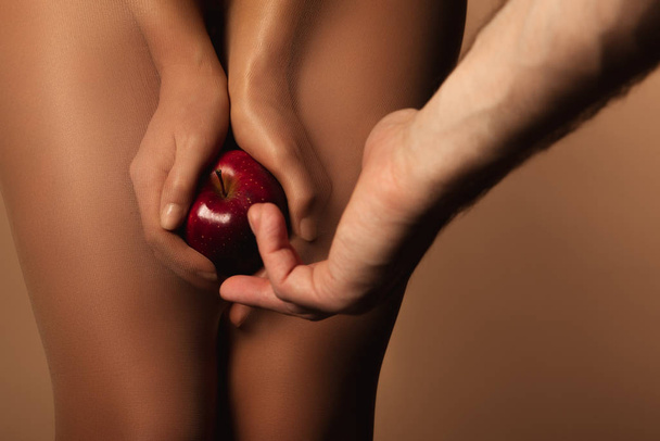 обрізаний вид чоловіка, який досягає жінки в нейлонових колготках, тримає стигле червоне яблуко ізольоване на коричневому
 - Фото, зображення