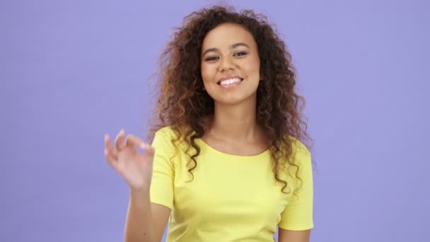 Beautiful happy african young woman in yellow t-shirt smiling and making ok gesture looking at the camera over purple background isolated - Video, Çekim