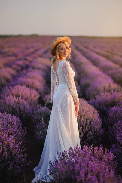 Young woman in white dress walks through blooming lavender field at sunset - Fotó, kép