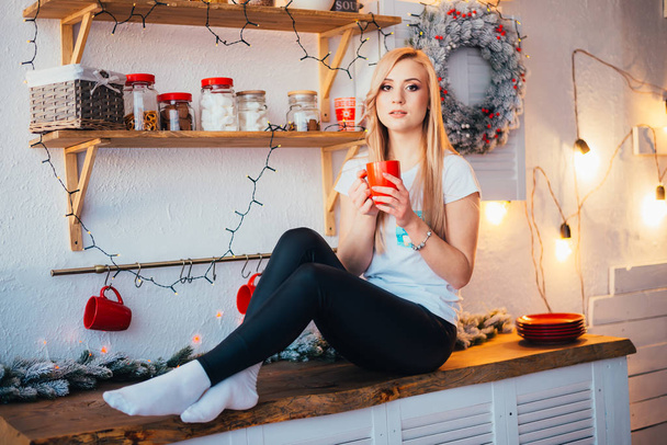 young blonde woman holding cup and looking at camera while sitting in kitchen at christmastime - Foto, imagen