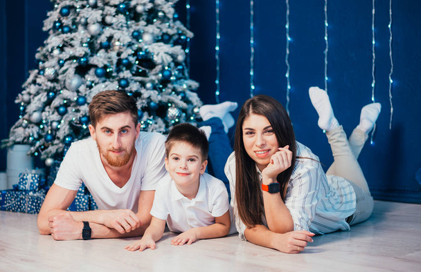 happy young family with adorable little son lying together and smiling at camera near christmas tree   - Photo, Image