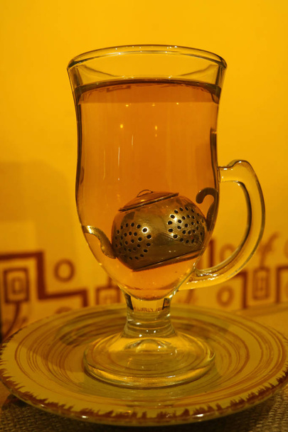 黄色の壁の前で提供されるかわいい茶葉フィルター付き透明ティーカップでホットカモミールティー - 写真・画像