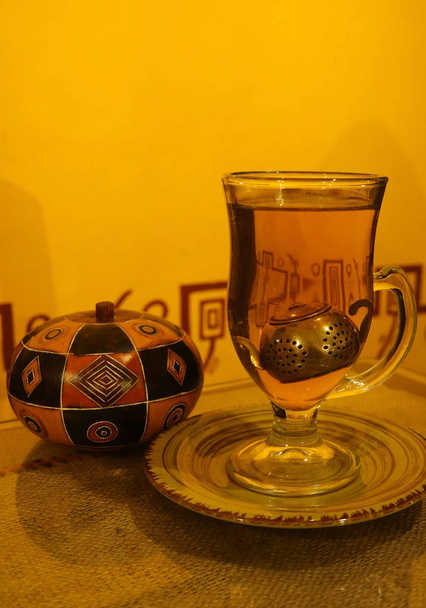 eine Tasse Kamille heißen Tee mit niedlichen Teeblatt-Filter in transparenter Tasse und peruanische hölzerne Zuckerkanne im Hintergrund - Foto, Bild