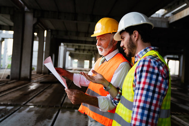 Two satisfied engineers, architects talking at building site with construction structure in background - 写真・画像