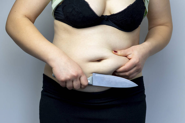 Woman in a black bra. Holds a knife near the abdomen. Close-up. On white background. The concept wants to cut excess weight - Photo, Image