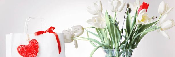Shopping bag, red heart and ribbon, gift and white bouquet tulips bouquet in a vase on white background. - Photo, Image