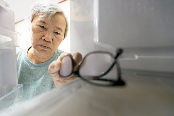 asiatische Seniorin mit Gedächtnisstörungen, vergisst ihre Gläser im Kühlschrank oder lagert sie im Kühlschrank, ältere Frauen mit Demenz, kognitiver Beeinträchtigung, Alzheimer, Amnesie - Foto, Bild