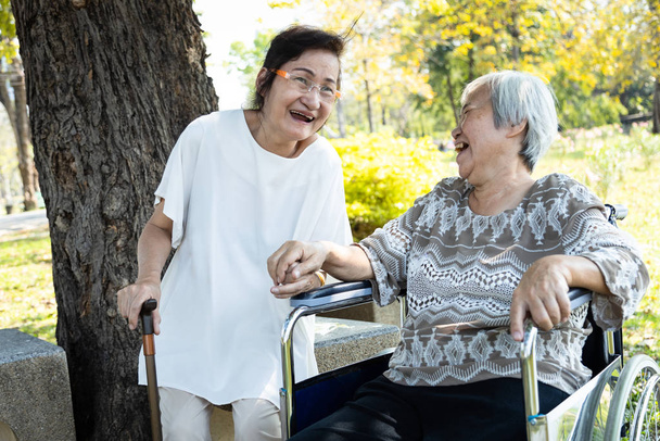 歩行棒を持つアジアの高齢者の女性は、車椅子で彼女のシニア親友と話を楽しみ、一緒に笑って、手を取り合って公園でリラックスしている2人の高齢者、子供の頃の友人、親友    - 写真・画像