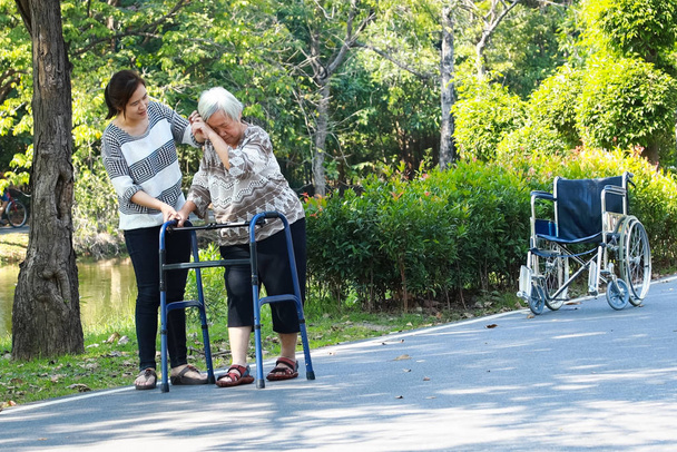 Asiático anciano uso de ayuda para caminar durante la rehabilitación después de la cirugía de rodilla, joven cuidador que ayuda a tranquilizar a las personas mayores práctica caminar con caminante se siente cansado, mujer cuidador ayuda, cuidado, apoyo
  - Foto, Imagen