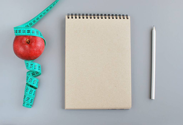 Plan de dieta, cuaderno vacío, lápiz, manzana roja y cinta métrica, tendido plano sobre fondo gris
. - Foto, imagen
