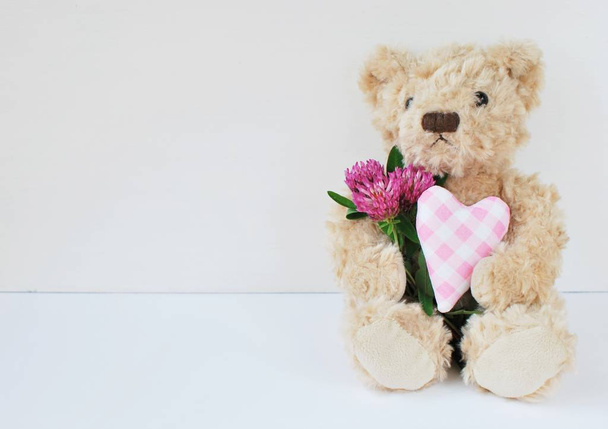 Cute soft toy teddy bear sitting on white background and holding purple heart and clover flower, place for text. - Photo, Image