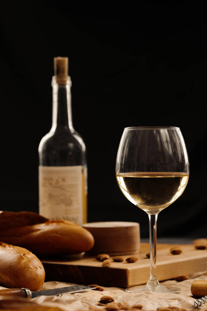 A glass of white wine on a table on a dark background. Baguette, cheese and nuts on the board - Фото, изображение