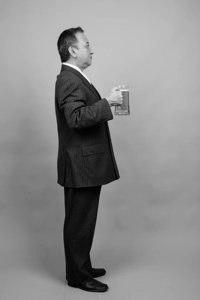 Студийный снимок зрелого азиатского бизнесмена, держащего стакан пива на сером фоне в черно-белом - Фото, изображение